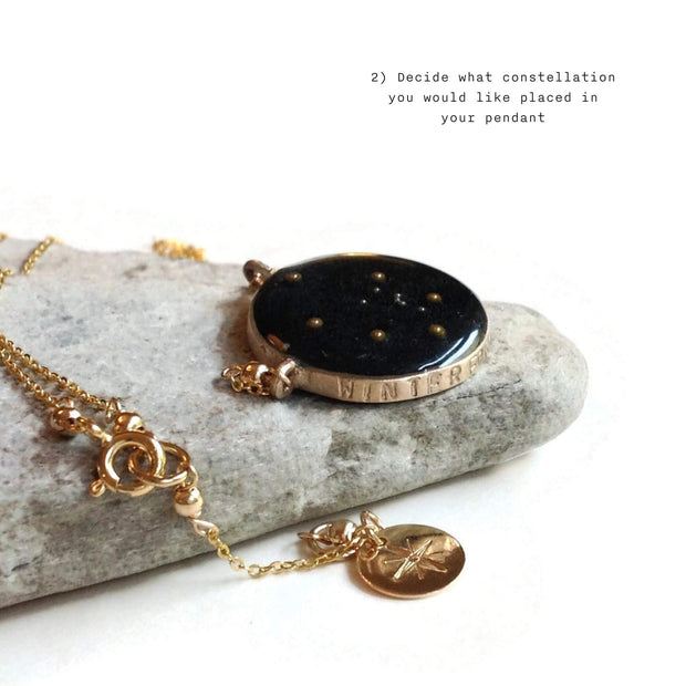Close up shot of a gold necklace with a pendant inlayed with a night sky colored resin and includes the winter hexagon stars in the bezel. Necklace is set on a rock with the chain falling over the front and a starburst stamped on a jewelry tag. Image has a white background. Text in the top right of the image says " 2) Decide what constellation you woudl like placed in your pendant"