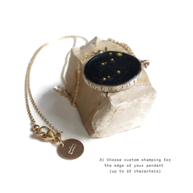 Necklace with a gold chain and bezel sitting on a sandstone rock. Bezel of necklace is inlayed with resin the color of night sky and the constellation Lyra formed from gold beads. Stamping along the band of the pendant says 10.02.2016. The chain of the necklace drapes around the front of the rock and has a jewelry tag with the libra symbol of the scaled stamped on it. The white background of the image has the text "3) Choose custom stamping for the edge of your pendant (up to 22 characters)"