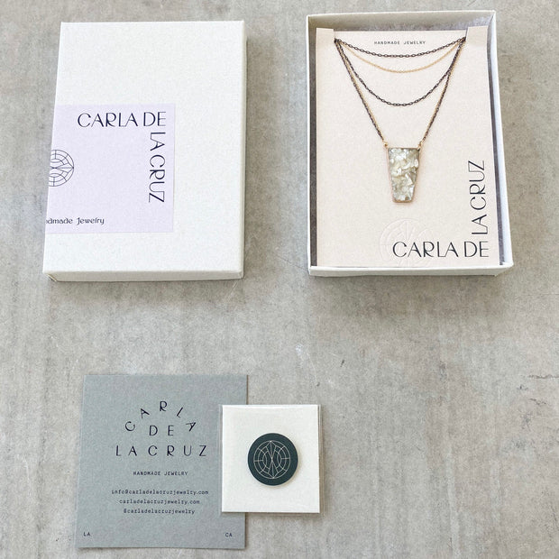 Five strand necklace shown in packagaing. Box is open showing the necklace inside. Sticker on box reads "Carla De La Cruz  Handmade Jewelry". Polising cloth and care card shown below packaging. Necklace chains alternate between oxidized silver and gold filled. A trapezoid pendant filled with crushed mother of pearl is attached to the two lowest chains of gold and silver. 