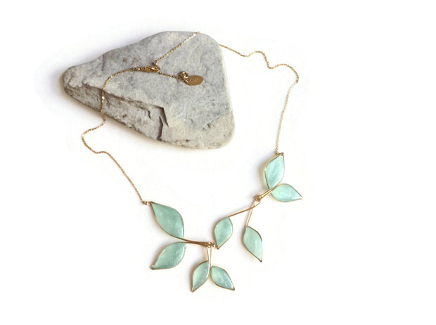 Gold necklace with 8 hanging leaf shaped pendants filled with clear sky blue resin with subtly  sparkling mica. Necklace is laying on a rock atop a white background, showing its adjustable length chain, clasp and CDLC jewelry tag. 