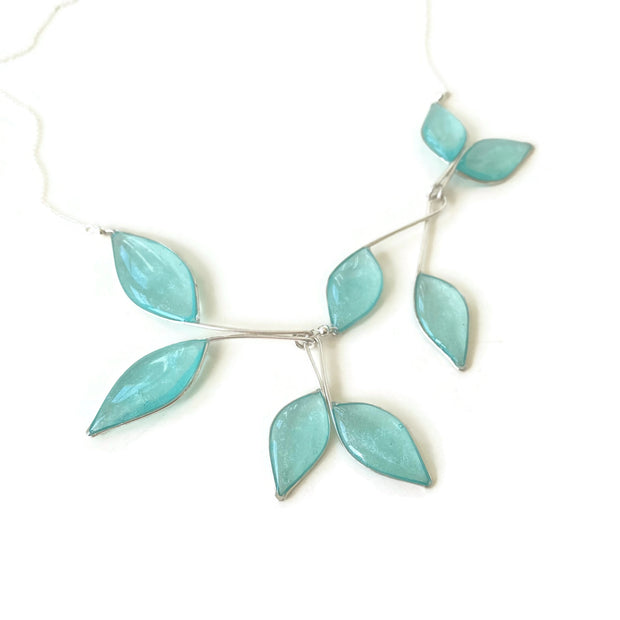 Silver Necklace with 8 hanging leaf shaped pendants filled with clear sky blue resin with subtly  sparkling mica. Necklace is laying flat on a white backgroun/ 