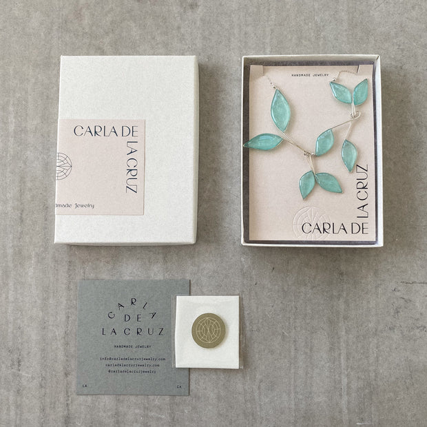 Silver Necklace with 8 hanging leaf shaped pendants filled with clear sky blue resin with subtly  sparkling mica. Necklace is shown in its packaging, a jewelry box with a sticker that says Carla De La Cruz, handmade jewelry. A care card and polishing cloth are displayed below the box. Concrete background. 