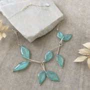 Silver Necklace with 8 hanging leaf shaped pendants filled with clear sky blue resin with subtly  sparkling mica. Necklace is laying on a broken piece of white marble set on a concrete background. Dried leaves peek into the edges of the photo. 