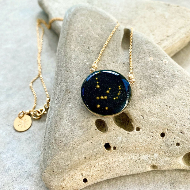 Image with a necklace placed on a stone with slate background. Gold filled necklace with night sky background and the Orion constellation made of gold beads. 