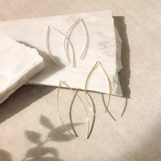 Gold and silver earrings in the shape of an leaf hoop sit on broken marble across concrete. The shadow of an olive branch covers the bottom corner of the image