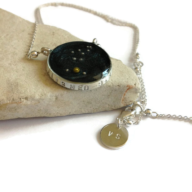 Image with a necklace placed on a stone with white background. Sterling silver necklace with night sky background and constellation made of gold beads. Stamping says “Aligned” across the bezel. Custom charm stamped with the initials “VS”