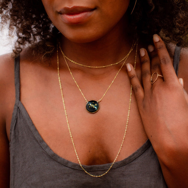 A woman with dark skin and dark curly hair wears a gold chain layered with a solar system necklace. She is wearing a gold open-circle ring, and her hand is placed on her shoulder. She is dressed in a grey tank dress with thin straps made of linen.