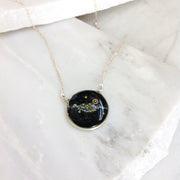 A silver Pendant necklace with the solar system made of small beads hovers over the milky way in dark sparkling background inside the necklace. It sits on pieces of broken marble. 